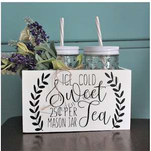 May include: White wooden box with a sign that reads "Ice Cold Sweet Tea 25¢ per Mason Jar". The box has two glass mason jars with white lids and straws in them. There are purple and white flowers in front of the box.