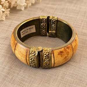 May include: A hinged bangle bracelet featuring curved, light brown, textured segments separated by two ornate, antique-gold-colored metal panels with embossed scrollwork. A white rectangular sticker inside reads MADE IN INDIA Item No. 11.