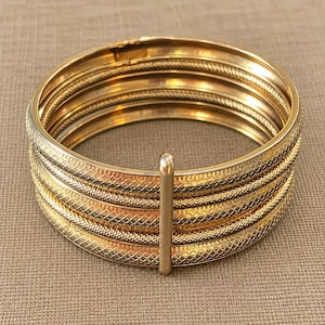 May include: A gold-tone hinged bangle bracelet featuring five stacked bands with a textured, cross-hatched pattern. The bands are connected by a single vertical metal bar on the front, and the bracelet includes a secure clasp mechanism.