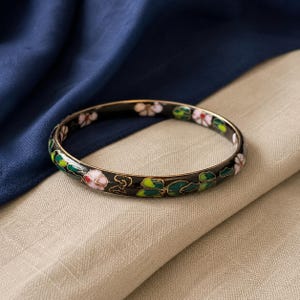 May include: A round bangle bracelet featuring a black base with gold-tone metal outlines. The surface is decorated with an enamel pattern of light pink flowers, green leaves, and small gold-colored cloud-like shapes arranged in a continuous design.