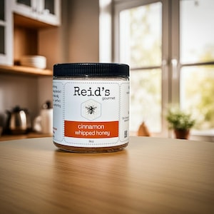 May include: A jar of Reid's Gourmet cinnamon whipped honey, 9oz, sits on a wooden table. The label features a bee illustration and the text "cinnamon whipped honey". The jar has a black lid and is in a kitchen setting.