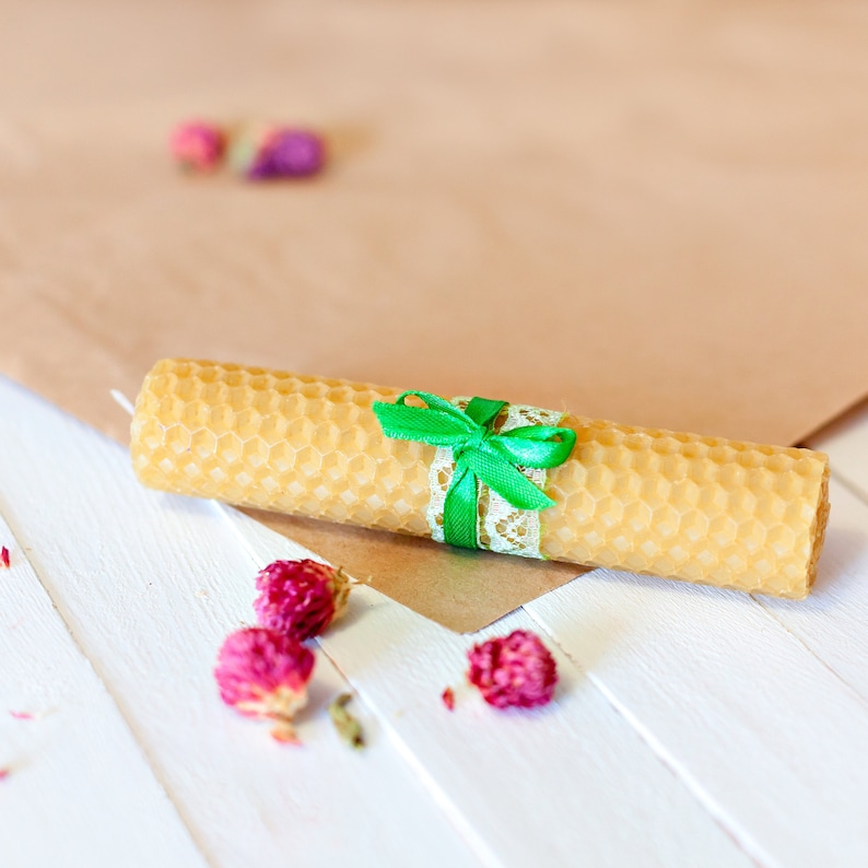 Pu&ograve; includere: Una candela arrotolata in cera d'api con un motivo a nido d'ape, legata con un nastro verde e pizzo. La candela poggia su una superficie di legno bianco, con boccioli di rosa secchi sparsi. Lo sfondo presenta carta marrone.