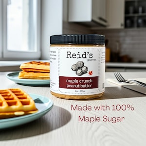 May include: A jar of Reid's gourmet maple crunch peanut butter, 255g, is displayed. The label shows a peanut illustration and the text "Made with 100% Maple Sugar." Waffles on blue plates are in the background.