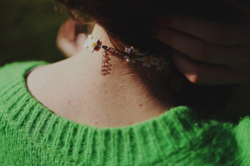 May include: A close-up of a person wearing a green jumper with a white daisy flower choker necklace.