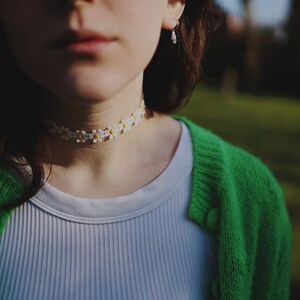 May include: A person wearing a white ribbed vest top with a green cardigan and a daisy flower choker necklace.