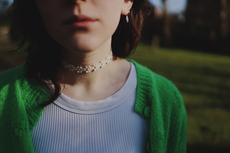 May include: A person wearing a white ribbed vest top with a green cardigan and a daisy flower choker necklace.