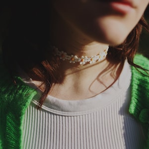 May include: A close-up of a person wearing a white shirt and a green cardigan. The person is wearing a white daisy flower choker necklace.
