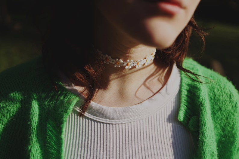May include: A close-up of a person wearing a white shirt and a green cardigan. The person is wearing a white daisy flower choker necklace.