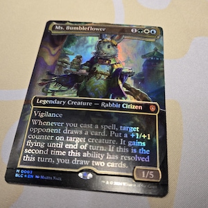 May include: A holographic Magic: The Gathering card featuring Ms. Bumbleflower, a Legendary Creature Rabbit Citizen. The card has a colorful illustration of a rabbit in a detailed environment. Text includes "Vigilance" and other game instructions.