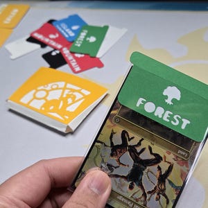 May include: A hand holding a card with a green "FOREST" label and a tree icon. Other cards with labels like "MOUNTAIN" and "EXILED" are visible. A yellow card with a design is also present, suggesting a card game theme.