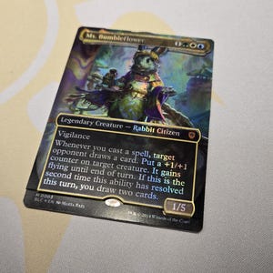 May include: A Magic: The Gathering card featuring "Ms. Bumbleflower," a Legendary Creature - Rabbit Citizen. The card has a colorful illustration of a rabbit-like figure. Text includes "Vigilance" and other game instructions. The card is marked 1/5.