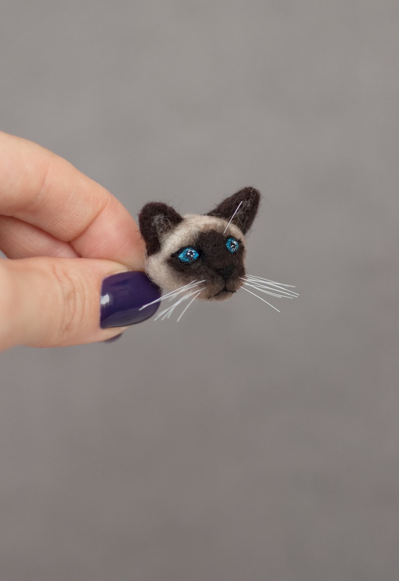 A woman's hand holds a cute miniature needle felted Siamese cat pin, side view. The pin is on gray background. Siamese cat brooch is made of wool. Siamese cat has blue eyes and cute white whiskers.