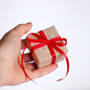 A woman's hand holds a small gift box made of brown craft cardboard, wrapped with twine and decorated with a red satin ribbon.
