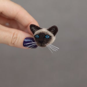 A woman's hand holds a cute miniature needle felted Siamese cat pin, side view. The pin is on gray background. Siamese cat brooch is made of wool. Siamese cat has blue eyes and cute white whiskers.