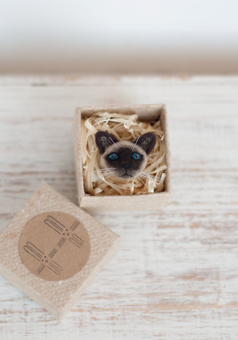 A cute miniature Siamese cat pin in the gift box. The gift box is made of brown craft cardboard.