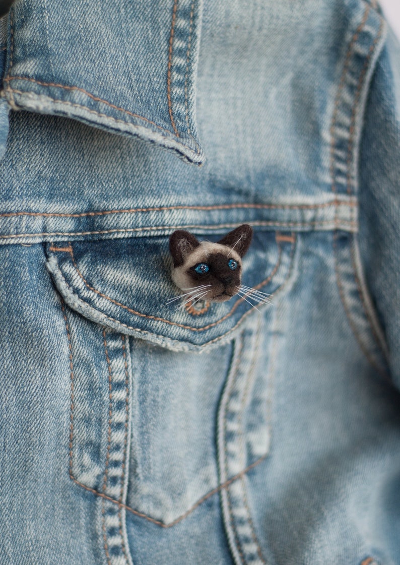 A cute miniature Siamese cat pin is attached to the flap of the blue denim jacket pocket, side view.