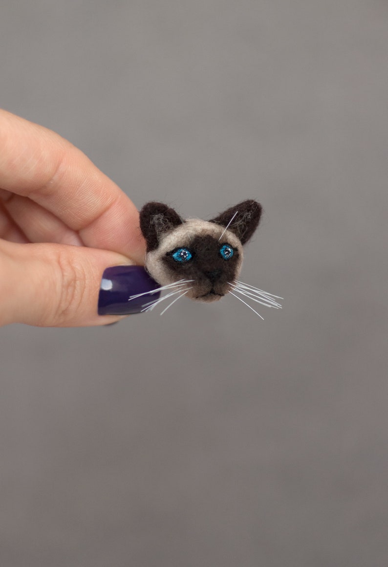 A woman's hand holds a cute miniature needle felted Siamese cat pin, side view. The pin is on gray background. Siamese cat brooch is made of wool. Siamese cat has blue eyes and cute white whiskers.
