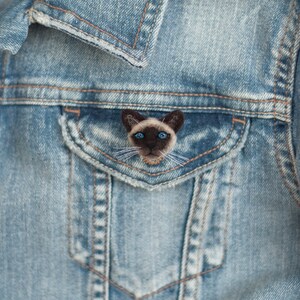 A cute miniature Siamese cat pin is attached to the flap of the blue denim jacket pocket.