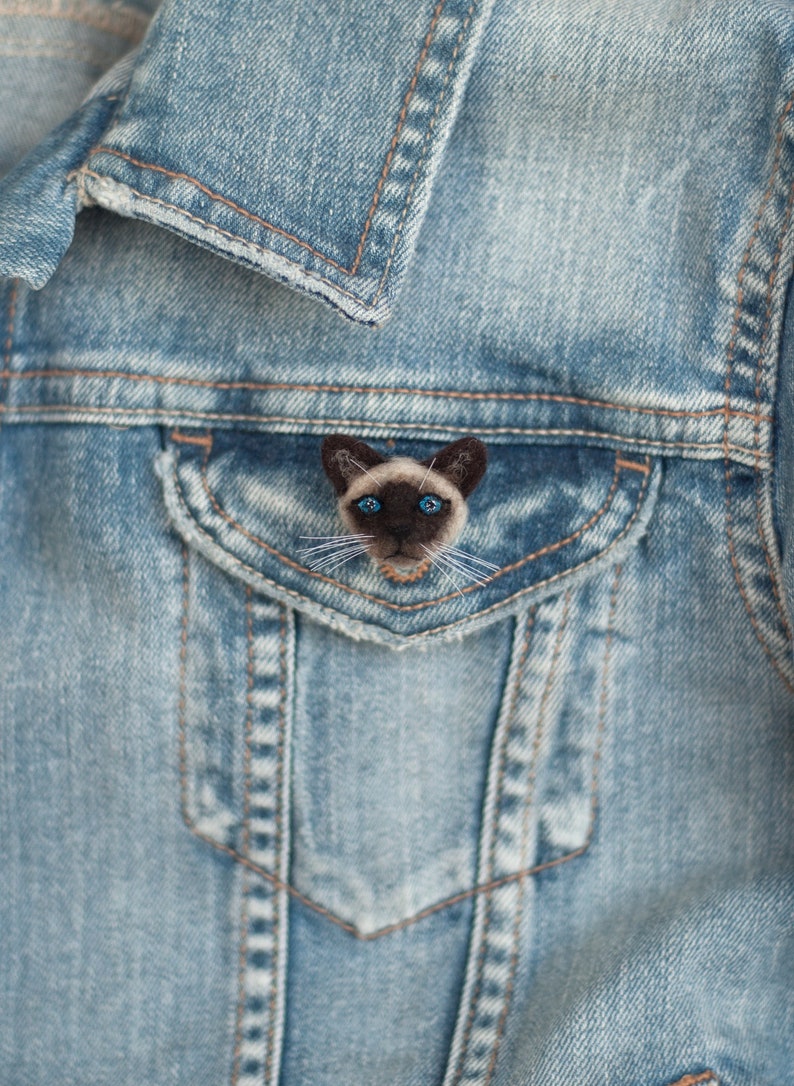 A cute miniature Siamese cat pin is attached to the flap of the blue denim jacket pocket.