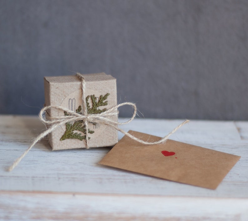 A woman's hand holds a small gift box made of brown craft cardboard, wrapped with twine and decorated with a green twig of thuja.