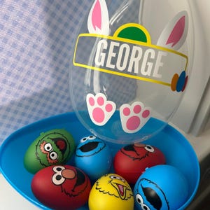 May include: A blue plastic Easter basket filled with character-themed eggs and a clear balloon. The balloon has bunny ears, pink paw prints, and the name "GEORGE" in yellow and white text. The eggs feature Sesame Street characters.