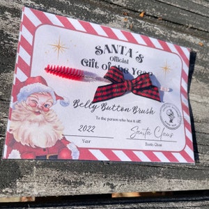 May include: A white card with red and white stripes around the border. The card has a red and black checkered bow, a red and white striped brush, and a cartoon image of Santa Claus. The card reads "SANTA'S Official Gift of the Year Belly Button Brush To the person who has it all 2022 Year Santa Claus Santa Claus".