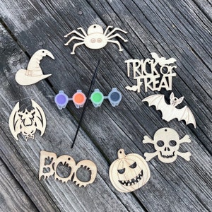 May include: A set of eight unfinished wooden Halloween ornaments with paint and a paintbrush. The ornaments include a witch's hat, a spider, a bat, a pumpkin, a skull and crossbones, a ghost, and two bats with the words "Trick or Treat".