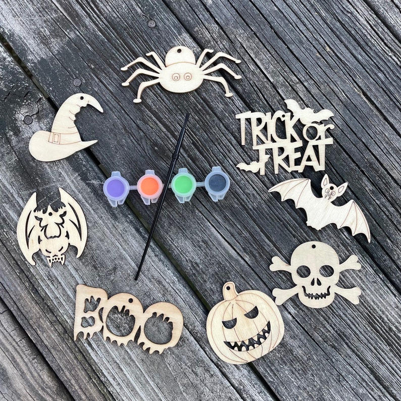 May include: A set of eight unfinished wooden Halloween ornaments with paint and a paintbrush. The ornaments include a witch's hat, a spider, a bat, a pumpkin, a skull and crossbones, a ghost, and two bats with the words "Trick or Treat".
