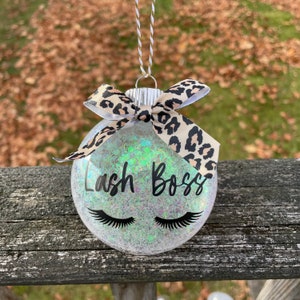 May include: A clear glass Christmas ornament with a glittery iridescent finish. It has a black and white leopard print ribbon tied around it and the words "Lash Boss" written in black with two black eyelashes underneath.