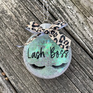 May include: A clear glass ornament with iridescent glitter and the words "Lash Boss" in black script. Black eyelashes are below the text. A leopard print bow and a gold and white string complete the decoration.