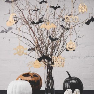 May include: A bare branch tree with black bats and Halloween-themed wooden ornaments hanging from it. The ornaments include a skull and crossbones, a spider, a pumpkin, a witch's hat, and a bat. The tree is in a clear glass vase and there are two pumpkins on the table below.