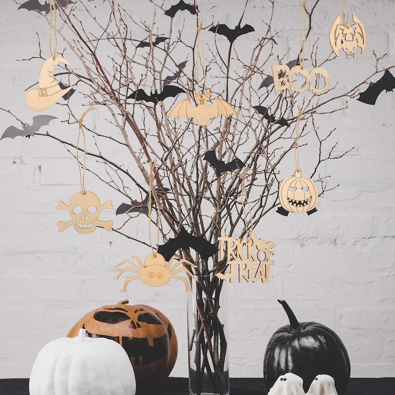 May include: A bare branch tree with black bats and Halloween-themed wooden ornaments hanging from it. The ornaments include a skull and crossbones, a spider, a pumpkin, a witch's hat, and a bat. The tree is in a clear glass vase and there are two pumpkins on the table below.