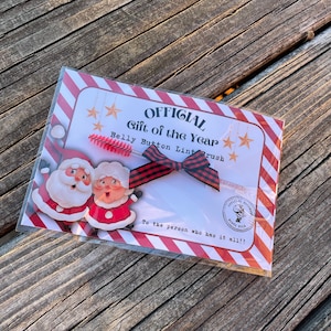 May include: A red and white striped card with the text "OFFICIAL Gift of the Year Belly Button Lint Brush" and a red and black bow. The card features a cartoon image of Santa Claus and Mrs. Claus. The text "To the person who has it all!!" is printed below the bow.