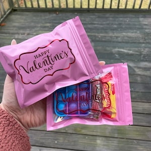 May include: Two pink zippered bags with the text "Happy Valentine's Day" printed on them. One bag contains a blue and purple pop-it toy, candy, and a Valentine's Day sticker.