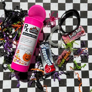 May include: A pink Halloween-themed prescription bottle with the text "Happy Halloween" and "You've been BOO'd!" is surrounded by candy, plastic spiders, and confetti on a black and white checkered surface.