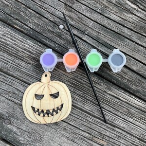 May include: A wooden pumpkin cutout with a carved face, ready to be painted. Four small paint pots with purple, orange, green, and black paint are included, along with a black paintbrush.