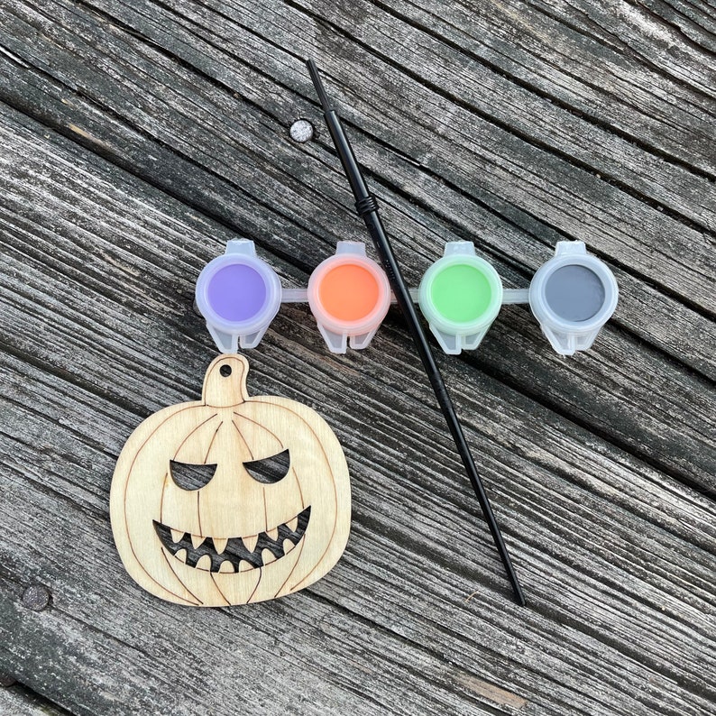 May include: A wooden pumpkin cutout with a carved face, ready to be painted. Four small paint pots with purple, orange, green, and black paint are included, along with a black paintbrush.