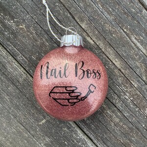 May include: A rose gold glitter Christmas ornament with the text "Nail Boss" and a black silhouette of a hand holding a nail file.