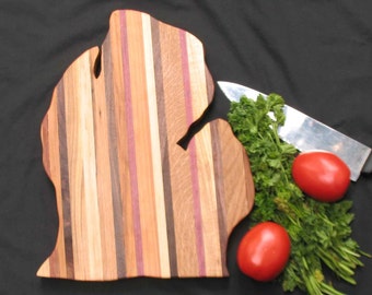 Puede incluir: Una tabla de cortar de madera con forma del estado de Michigan. La tabla está hecha de varios tipos de madera, creando un patrón a rayas. La tabla está rodeada de perejil y dos tomates rojos.