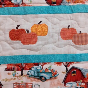 May include: A quilted fabric with a fall theme. The design features pumpkins, vintage trucks, and red barns set against a backdrop of autumn foliage. The quilt has a white background with teal borders and a quilted pattern.