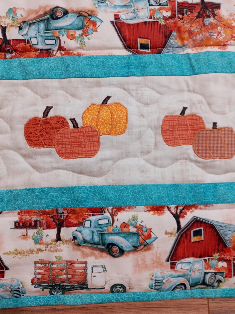 May include: A quilted fabric with a fall theme. The design features pumpkins, vintage trucks, and red barns set against a backdrop of autumn foliage. The quilt has a white background with teal borders and a quilted pattern.
