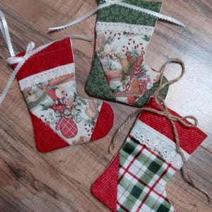 May include: Three small Christmas stockings made of fabric with different patterns. The stockings are red, green, and white with a white lace trim. The stockings are hanging on a wooden surface.