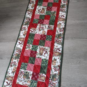 May include: A rectangular quilted table runner featuring a central patchwork design of red and green squares surrounded by a border of white fabric printed with snowmen, sleds, and evergreen trees, finished with a dark green fabric binding.