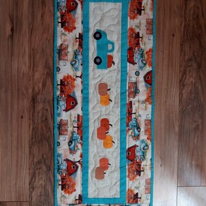 May include: A quilted table runner with a fall harvest theme. The runner features a central panel with a blue truck and appliqued pumpkins. The border has a repeating pattern of barns, trucks, and trees in autumn colors. The runner is approximately 12 inches wide and 36 inches long.