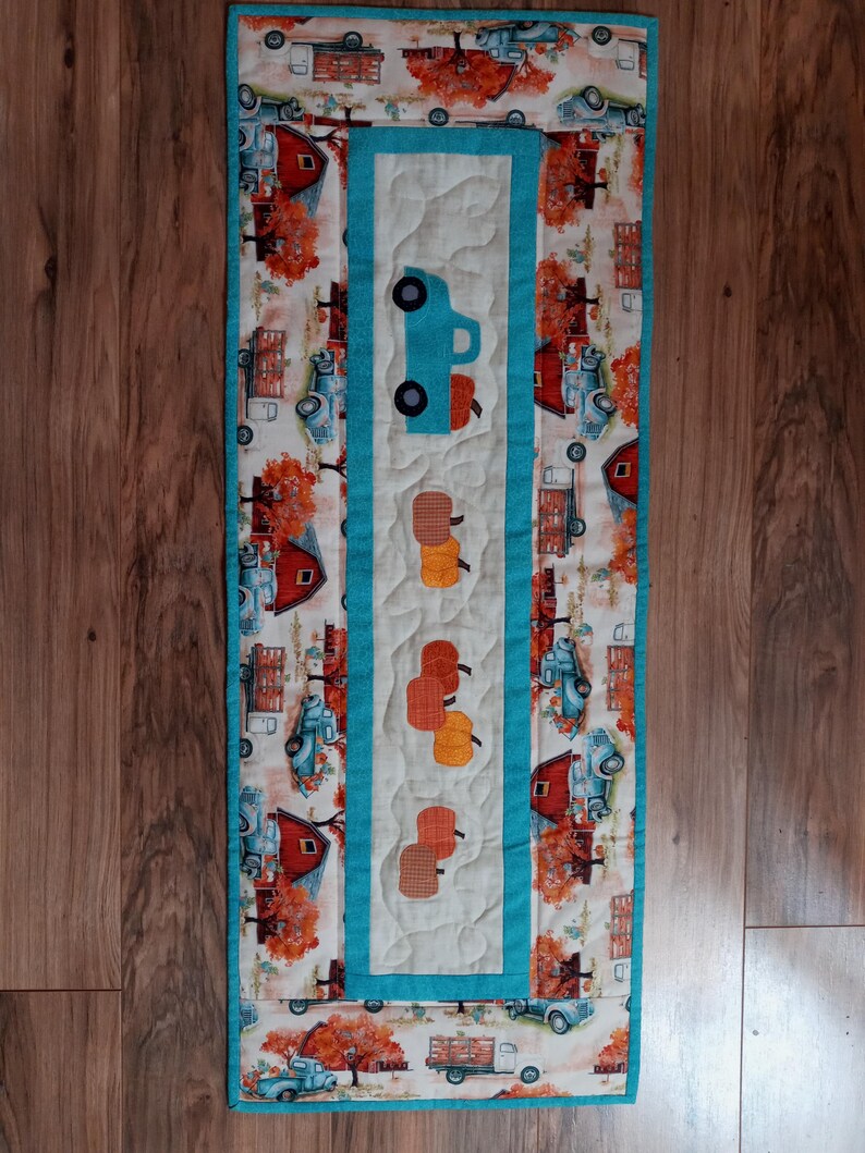 May include: A quilted table runner with a fall harvest theme. The runner features a central panel with a blue truck and appliqued pumpkins. The border has a repeating pattern of barns, trucks, and trees in autumn colors. The runner is approximately 12 inches wide and 36 inches long.