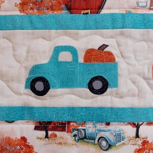 May include: A quilted fabric featuring a repeating pattern of vintage trucks carrying pumpkins. The trucks are in shades of blue, and the pumpkins are orange. The background includes autumn scenes with trees and barns.