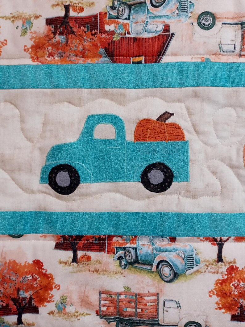 May include: A quilted fabric featuring a repeating pattern of vintage trucks carrying pumpkins. The trucks are in shades of blue, and the pumpkins are orange. The background includes autumn scenes with trees and barns.
