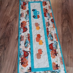 May include: A rectangular quilted table runner featuring a repeating pattern of vintage trucks, barns, and fall foliage. The center panel has a teal border and features a teal truck and several orange pumpkins. The runner is approximately 12 inches wide and 36 inches long.