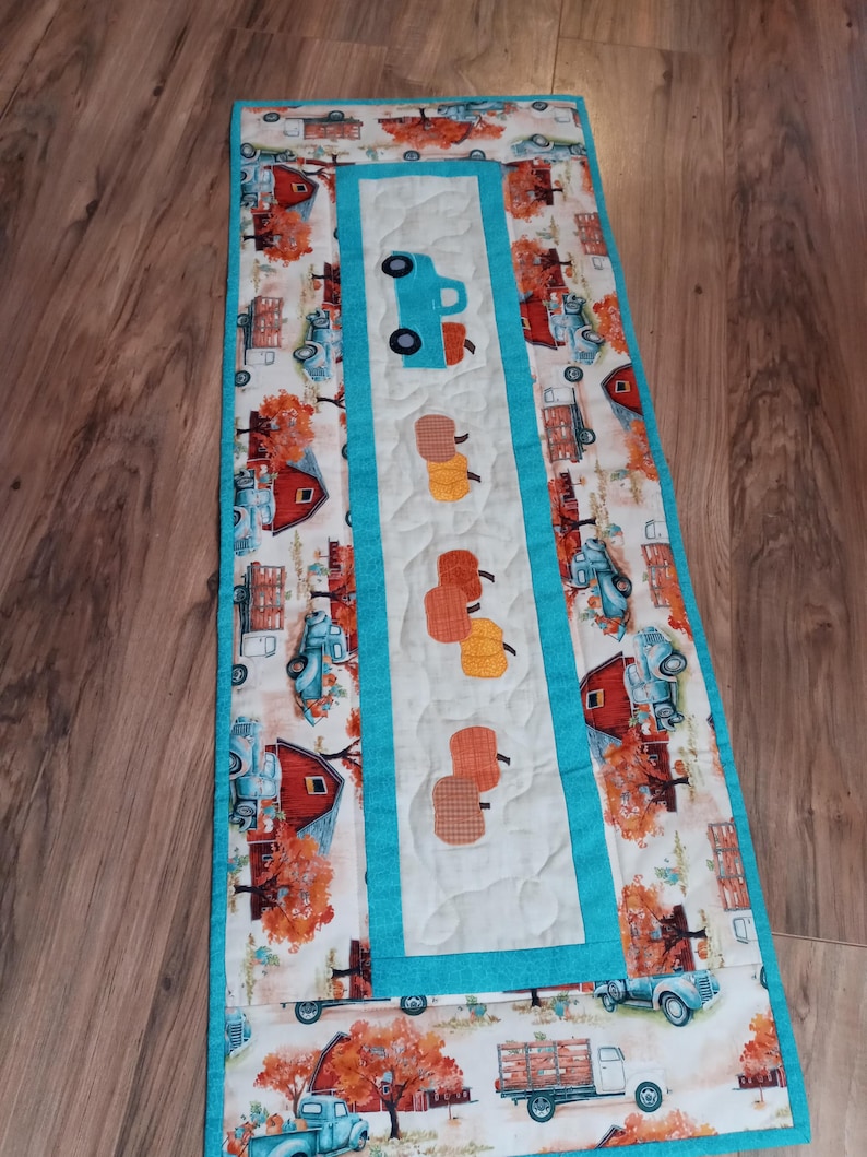 May include: A rectangular quilted table runner featuring a repeating pattern of vintage trucks, barns, and fall foliage. The center panel has a teal border and features a teal truck and several orange pumpkins. The runner is approximately 12 inches wide and 36 inches long.
