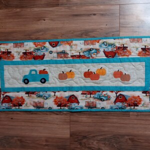 May include: A rectangular quilted table runner with a fall harvest theme. The runner features a central panel with a blue truck carrying pumpkins and several pumpkin appliqu&eacute;s. The border has a repeating pattern of trucks, barns, and fall foliage.